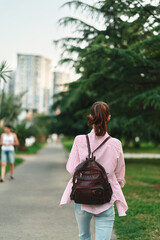 Naklejka premium Casual woman with backpack walks along a tree lined park path, city skyline in the distance, relaxed lifestyle mood, everyday outdoor exploration and gentle breeze on a sunny day