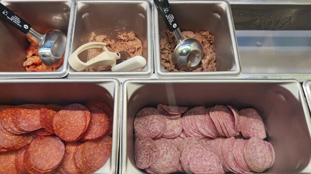 Variety of fresh ingredients for making sandwiches displayed in stainless steel trays. Deli counter showing sliced salami and meat filling