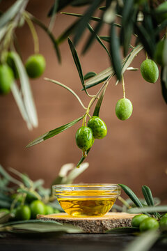 Small glass bowl filled with olive oil placed on wooden surface surrounded by fresh green olives and olive branches. Concept of Mediterranean cuisine, organic farming, healthy food and natural harvest
