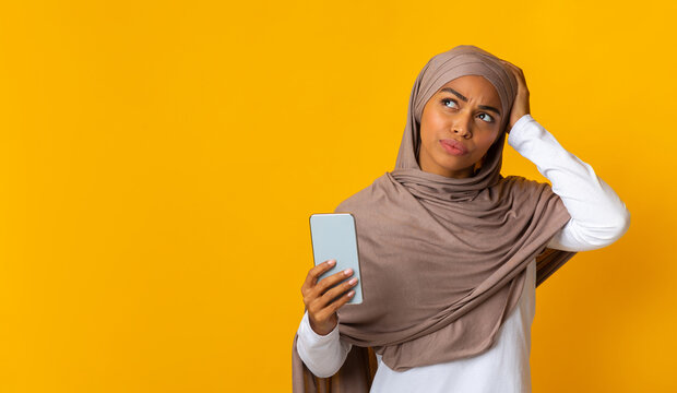 Forgot phone password. Thoughtful African American muslim woman holding smartphone and touching her head, not able to unblock her device, yellow background