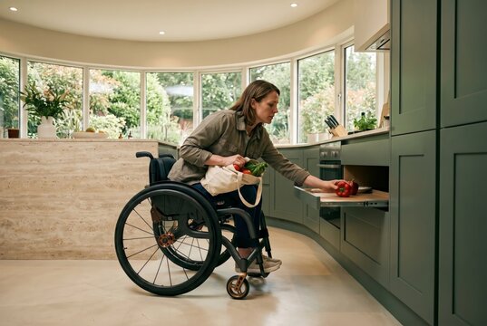Woman in modern wheelchair putting away groceries in lowered kitchen cabinets, accessible independent living