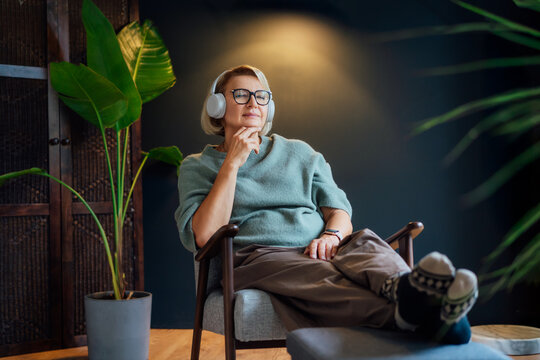 Middle aged mature woman with closed eyes enjoys music or meditation with headphones in hygge interior home with green plants. Take a break and relax. Lady enjoy calm mood, well being alone.