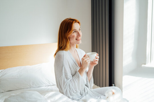 Relaxed and calm middle aged woman with cup of coffee relaxing in bed in morning sunlight. Hot drink for wake up and energy boost. Well being morning routine and daydreaming mood. Good morning habits
