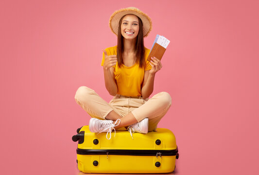 Emotional young woman in summer hat holding passport and plane tickets, sitting on big yellow luggage, ready to travel. Cheerful lady traveller posing over pink studio background, ready for vacation