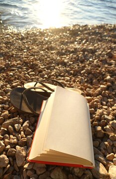 Book and flip flop sandals on the beach with pebbles in sunset by the sea, summer accessories background