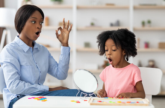 Pretty little girl attending speech therapy session at clinic, looking at mirror, beautiful young african american woman speech-language pathologist working on sounds with kid, articulating