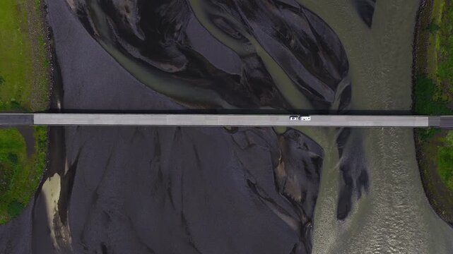 Aerial top down scene in southern Iceland shows a long bridge as a white car and camper move across, with pale grey meltwater weaving around black volcanic sand.