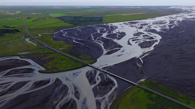 Aerial view shows milky glacial channels in black sand, Ring Road bridge, green farmland, a passing vehicle, and a flat coast in soft overcast daylight in Iceland.