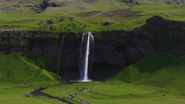 Aerial view shows Seljalandsfoss waterfall and a thin stream over basalt into a misty pool. Summer light reveals river, bridge, footpath, and tourists moving behind the falls.
