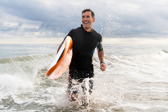 Middle aged sportive handsome man learning surfing and running in the waves holding board