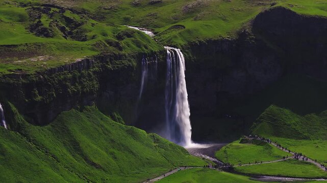Aerial view of Seljalandsfoss waterfall on Iceland south coast shows a single drop, mist, a plunge pool, a braided stream, tourists on paths, and a small footbridge.