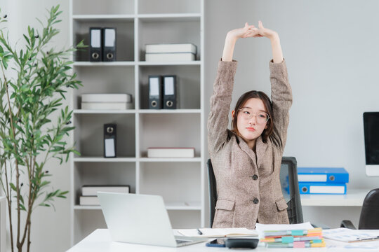 Woman Stretching at Desk: A young businesswoman in a professional setting stretches her arms overhead while sitting at her desk, indicating a moment of relaxation or a break from work.