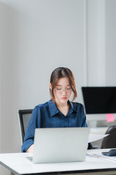 Focused Professional: A young woman in a blue shirt concentrates on her laptop at a modern office desk, surrounded by professional equipment.