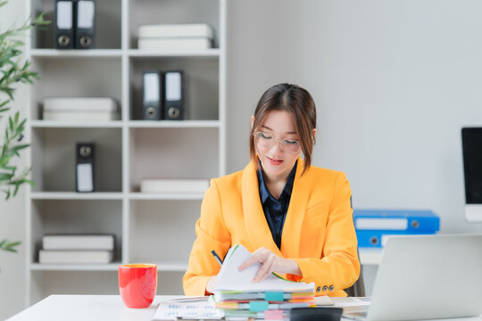 Focused Accountant at Desk: A young businesswoman in a vibrant yellow blazer meticulously reviews documents at her office desk, surrounded by professional supplies and a minimalist shelf.