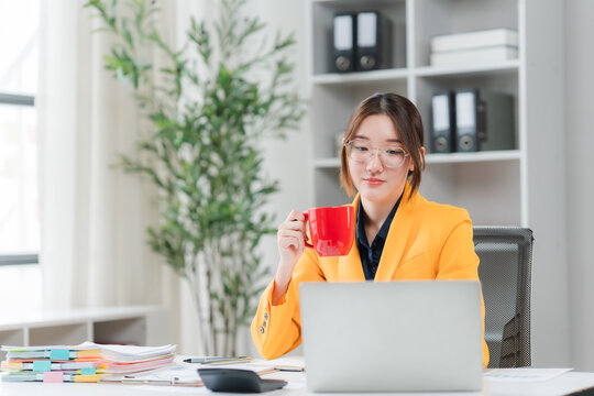 Morning Coffee Break: A young woman in a vibrant yellow blazer takes a moment to enjoy her coffee while working on her laptop in a bright, modern office setting.