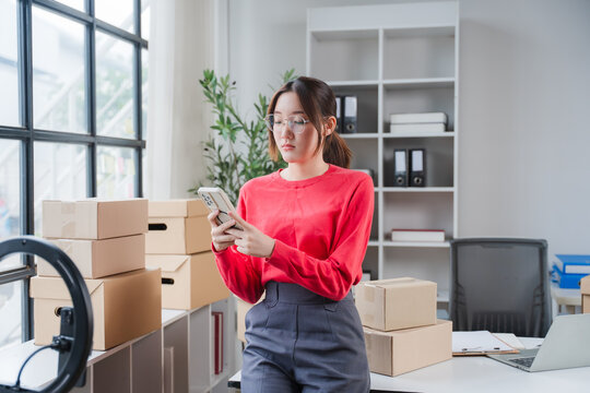 Concerned Woman in Office with Boxes: A young woman, appearing stressed or thoughtful, checks her phone amidst stacks of cardboard boxes in a home office or small business setting.