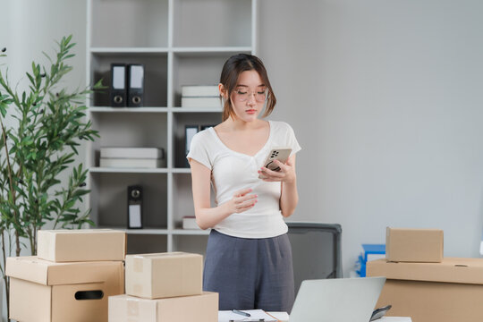 Entrepreneur's Workspace: A young woman with glasses focuses on her smartphone, surrounded by boxes and a laptop in a modern workspace, suggesting e-commerce or small business operations.