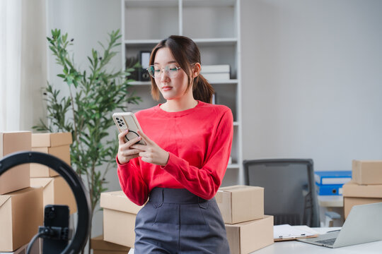 Young Entrepreneur Using Smartphone in Home Office: A focused young woman stands amidst shipping boxes, using her smartphone to manage her e-commerce business operations efficiently.