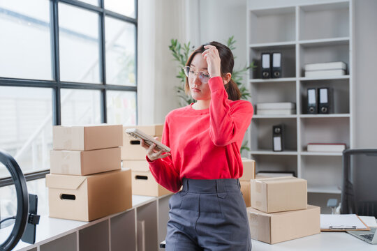 Frustrated Entrepreneurship: A young woman, surrounded by delivery boxes, looks stressed while holding a tablet, symbolizing the challenges of online business and e-commerce.