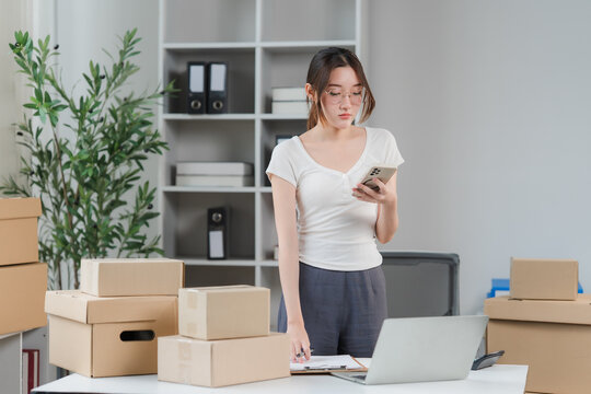 Businesswoman managing online order: A young businesswoman is diligently managing online orders in her home office, surrounded by boxes and a laptop, highlighting the entrepreneurial spirit.