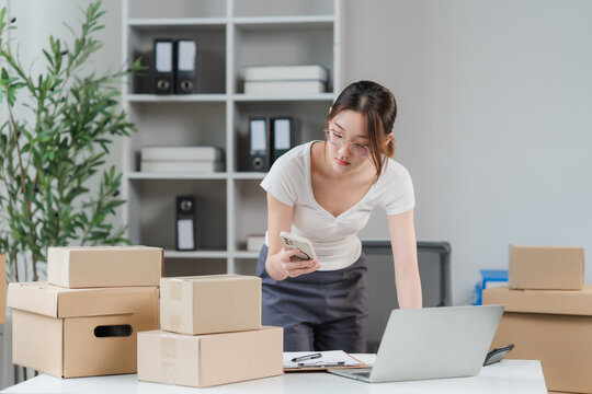 Young Woman Entrepreneur Managing Online Orders from Home Office, surrounded by shipping boxes and using a laptop and phone for her e-commerce business.