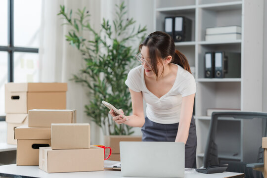 Startup Entrepreneur Organizing Orders: A focused female entrepreneur diligently checks orders on her phone amidst a desk laden with cardboard boxes and a laptop.