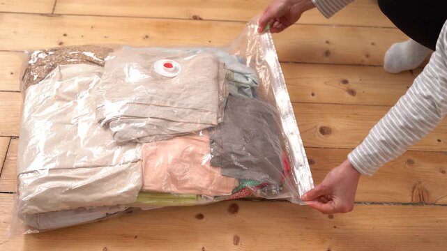A person's hands use a green slider to seal a clear vacuum storage bag filled with folded clothes on a wooden floor