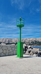 Fototapeta premium A vibrant green navigation lighthouse tower standing on a concrete pier in Baska on Krk island with mountains in the background.