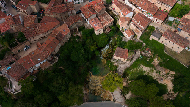 peaceful aerial view of a village with red-tiled roofs and stone buildings surrounded by greenery and a waterfall in a serene landscape setting with paved streets and parked cars.
