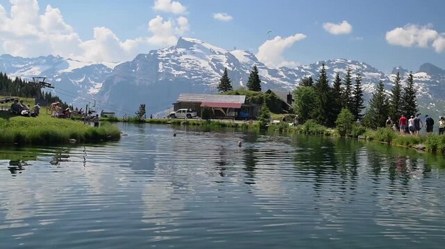 Engelberg, Switzerland - 27.06.2026: Hikers rest beside a reflective mountain lake with chalet and snowcapped peaks on a sunny summer day.