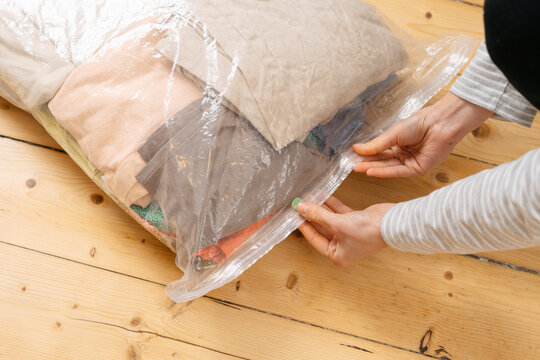 Female hands seal a transparent vacuum bag full of clothes. She organizes and compresses them for efficient storage or travel. The scene is on a light wooden floor with natural light
