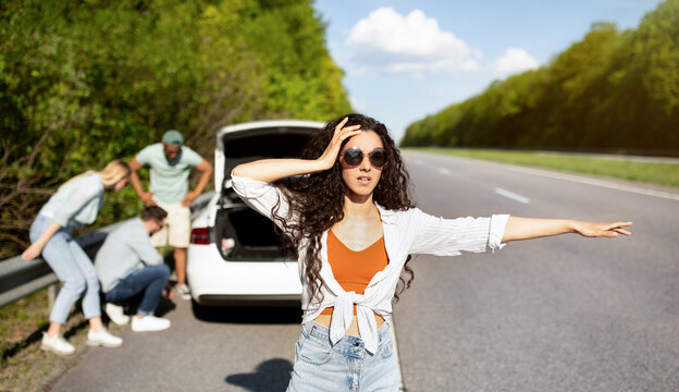 Millennial lady trying to stop passing car, asking for help, her friends fixing broken vehicle, having road accident. Young woman hitchhiking on highway, needing emergency assistance