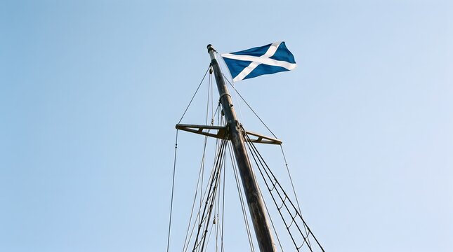 A Scottish flag fluttering majestically atop a tall mast against a clear blue sky. The iconic St Andrew's Cross design is prominent, symbolizing national pride and heritage