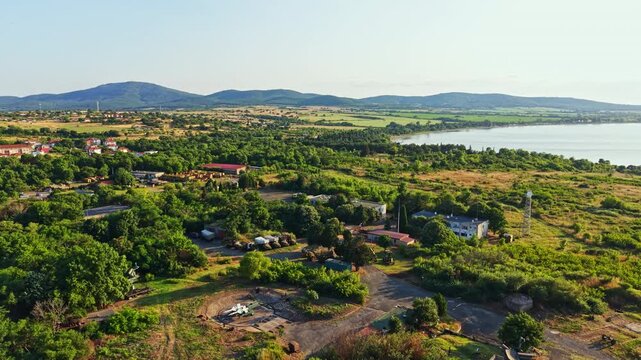 The scene shows an aerial view of a green area close to a lake. Houses and trees fill the landscape. Hills are visible in the background under clear skies.