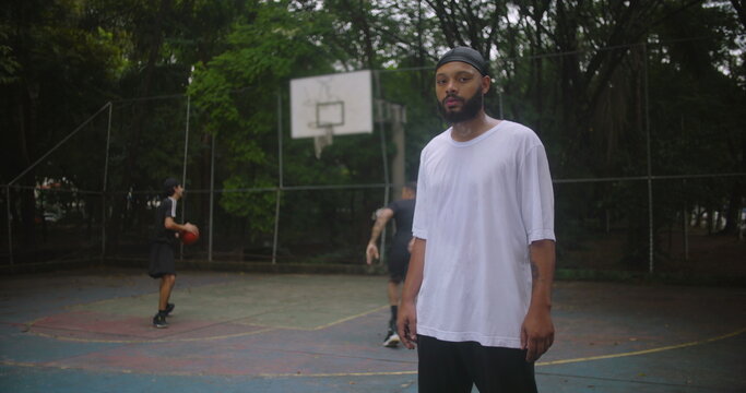 African descent man wearing durag standing on basketball court observing teammates play, showing calmness, confidence, and connection to the team during a casual urban game