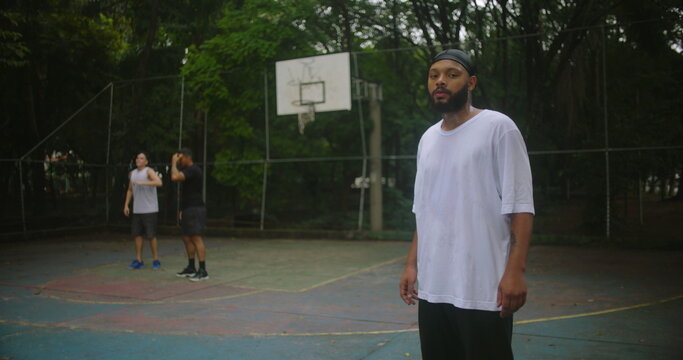 African descent man wearing durag with arms crossed on basketball court looking confident and focused while friends play behind, symbolizing leadership, resilience, and strength in teamwork