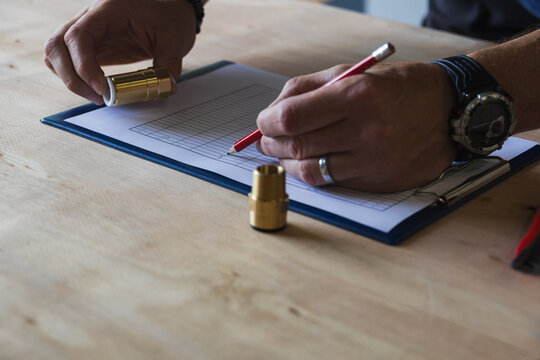 High angle shot of a worker writing with a red pencil on a clipboard chart next to brass components on a wooden table. Quality control