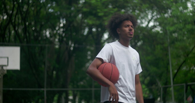 African American man standing on outdoor basketball court holding ball, thoughtful and apprehensive expression, reflecting quietly surrounded by trees
