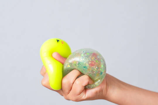 Colorful squishy toys held in child's hand, featuring a bright yellow squishy and a glittery, translucent slime-like toy against a plain background