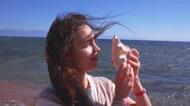 Happy young woman listening to the sound of the ocean in a seashell on a sunny beach