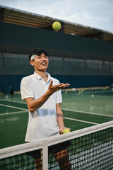 Obraz premium Young Asian Man Smiling and Tossing Tennis Ball at Net on Outdoor Court