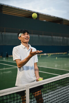 Young Asian Man Smiling and Tossing Tennis Ball at Net on Outdoor Court