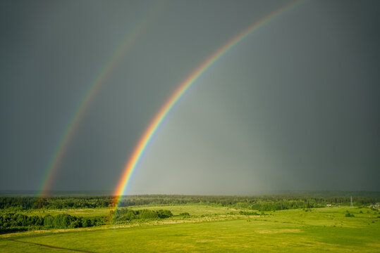 Double rainbow spanning a wide green landscape under a stormy sky, emerging after rainfall, creating a striking contrast between colorful light and gray heavens