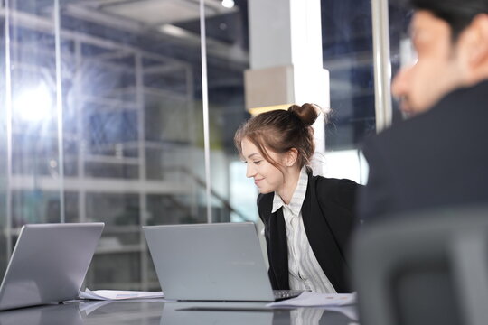 Businesswoman with a bun smiles gently while typing on her laptop as a dark-haired colleague leans in from the foreground in a modern glass-walled coworking office in Bangkok.