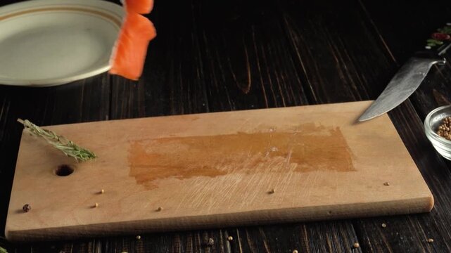 A salmon fillet is transferred from a wooden cutting board to a bowl, with herbs and kitchen utensils arranged around it, demonstrating the cooking process in a rustic kitchen environment