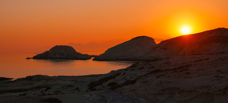 Golden morning sunrise over the rocky coastline of Milos near Mandrakia and Sarakiniko Beach, with the tranquil Aegean Sea and creating a serene, warm glow across the island&rsquo;s volcanic landscape.
