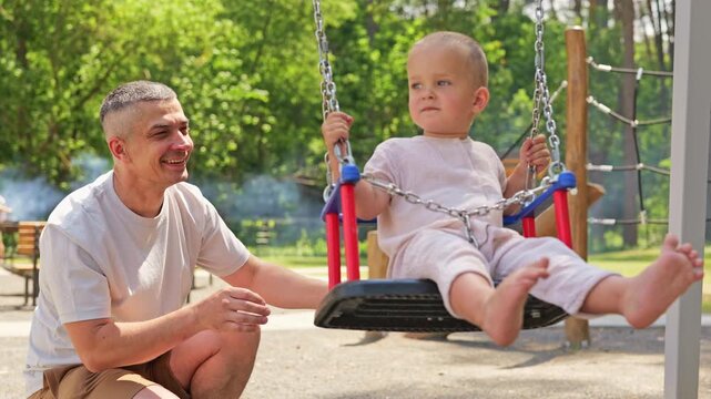 A man pushes his child on a swing at a playground. Nearby, other families are enjoying the park on a warm day. Laughter and sounds of play fill the air