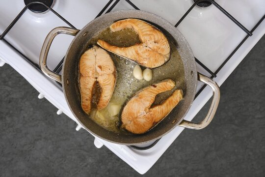 Salmon fillets are being cooked in a pan with garlic on a stovetop. The kitchen is well-lit and the fillets show a golden color, indicating they are almost ready to be served