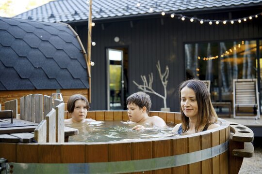 Three family members are immersed in a wooden hot tub outside a contemporary house during the evening. They are engaged in conversation, enjoying their time together in a cozy atmosphere