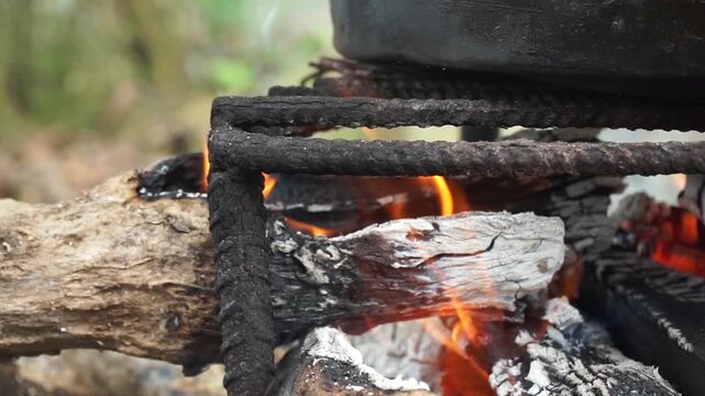 Traditional outdoor cooking over an open firewood campfire with bright orange flames under a large black metal pot on a rustic iron grill structure.